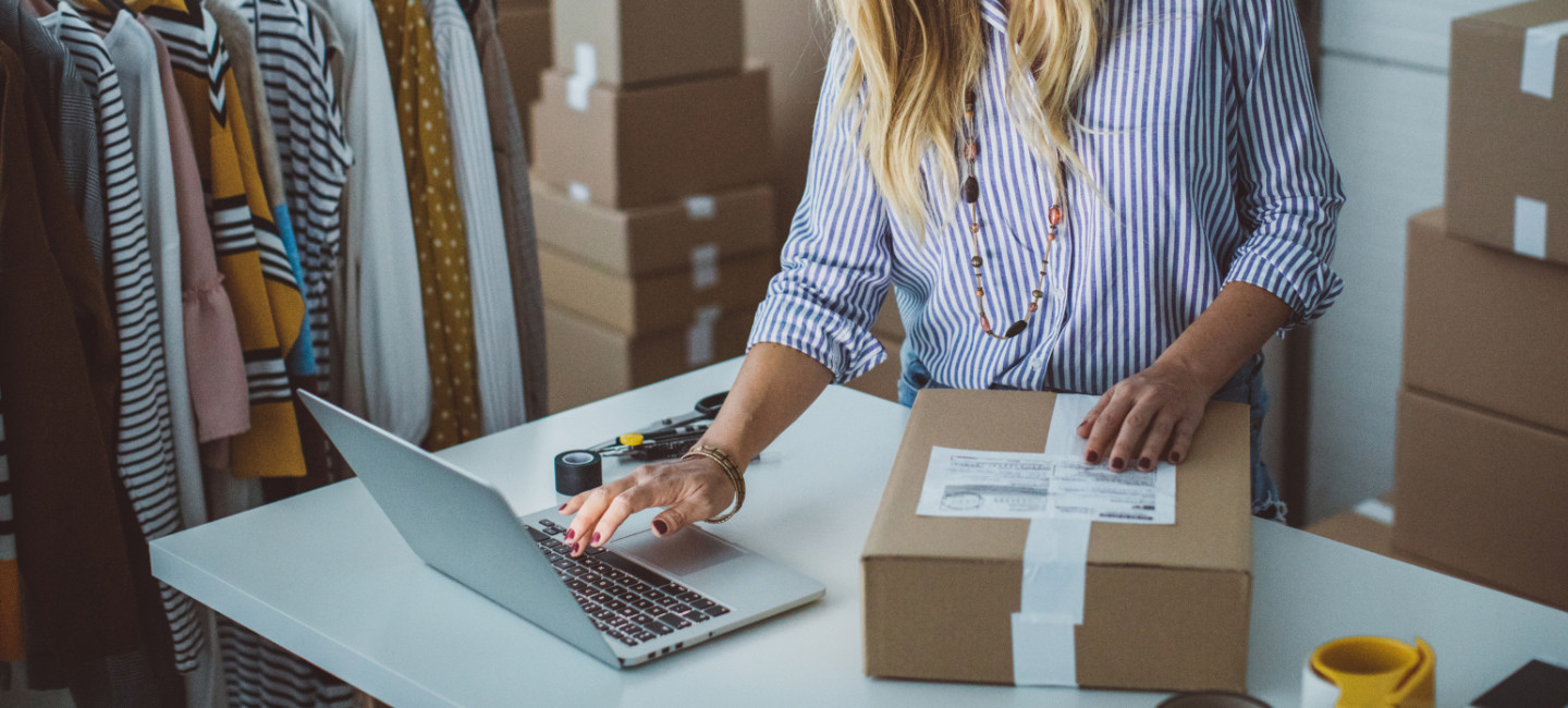 senhora num portátil  e com uma caixa com encomenda e rótulo. Várias roupas penduradas num charriot