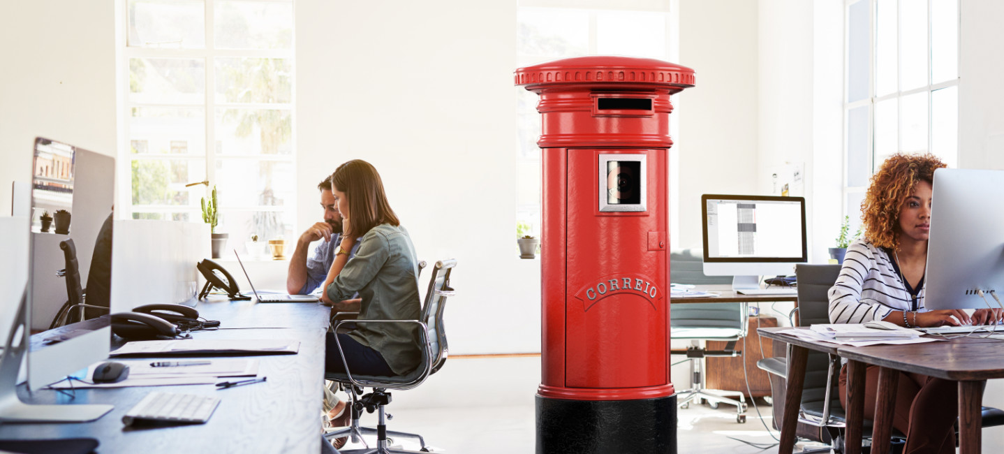 Ambiente de escritório com pessoas a trabalhar e um marco de correio vermelho no centro da sala.