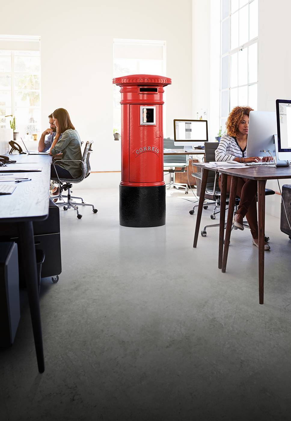 Ambiente de escritório com pessoas a trabalhar e um marco de correio vermelho no centro da sala.