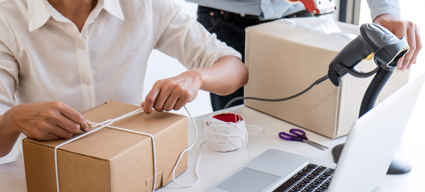 Duas pessoas embalam caixas com computador, scanner, tesoura e outros itens em cima da mesa.