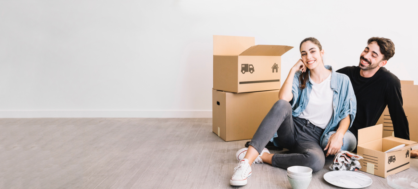 Um casal sentado no chão entre caixas de mudança, com itens domésticos à sua volta.